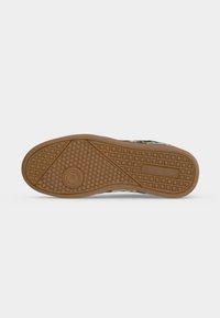 Semelle de chaussure avec caoutchouc marron, motif circulaire à l’avant, texture en grille au talon et au médio-pied, sur un fond clair uni.
