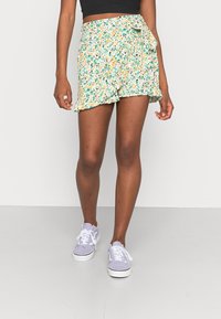 Miniwrap-kjol med blommönster på en mintgrön bakgrund, gul och teal blomma, volangkant och knytning i sidan.