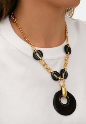 Collier en chaîne dorée présentant des maillons circulaires noirs alternés et un grand pendentif noir avec une découpe au centre.