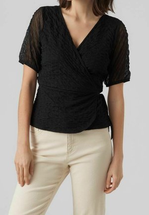 Femme portant un haut cache-cœur texturé noir avec des manches courtes transparentes et un pantalon couleur crème sur un fond uni.