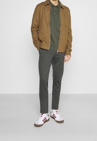 Veste zippée marron, polo vert, pantalon gris et baskets blanches avec des rayures rouges. Le matériau semble doux avec une finition texturée.