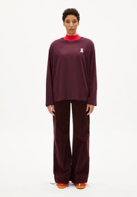 Langsleeve maroon shirt met een witte strikdetail, gecombineerd met bijpassende wijde corduroy broeken en oranje schoenen.