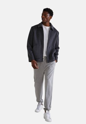 Veste en mélange de laine grise avec un col, deux poches avant, portée sur une chemise blanche et assortie à un pantalon de jogging gris et des baskets blanches.