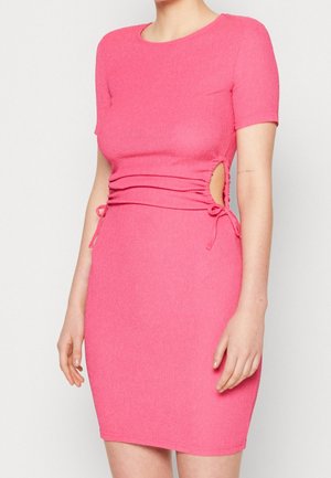 Woman wearing a fitted short-sleeve pink dress with side cutouts and tie details on a plain light background.