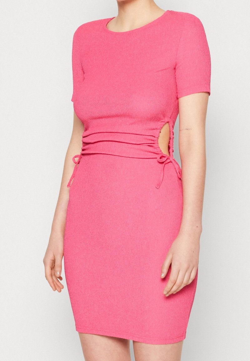 Femme portant une robe rose courte ajustée à manches courtes avec des découpes latérales et des détails à nouer sur un fond clair uni.
