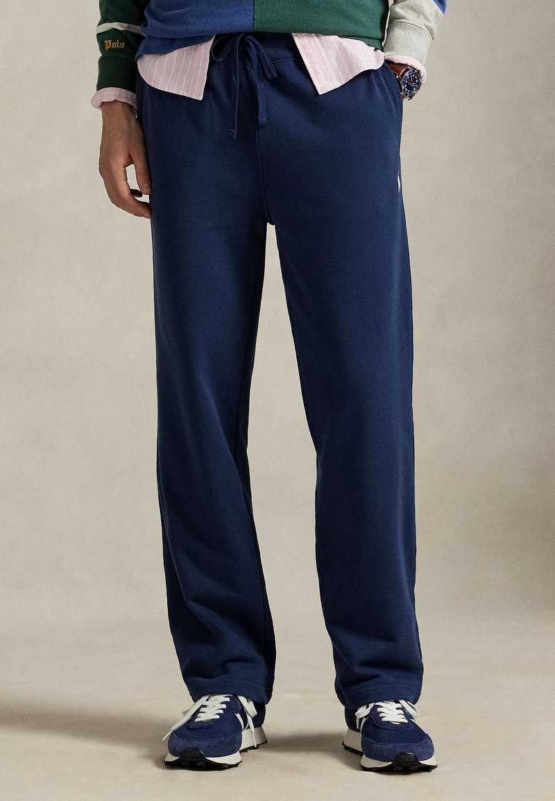 Tamsiai mėlynos medvilninės laisvalaikio kelnės, pagamintos iš minkštos medžiagos, su raišteliu juosmens srityje, šoninėmis kišenėmis ir laisvu siluetu, derinamos su sportiniais bateliais.