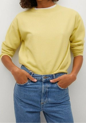 Osoba ubrana w żółty sweter o fakturze z białą lamówką oraz niebieskie, wysokowalne dżinsy, z rękami schowanymi w przednich kieszeniach.