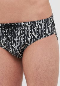 Slipuri de baie negre cu un design repetat de text alb și gri. Au un talie cu șnur și o croială strânsă. Fabrică netedă și elastică.