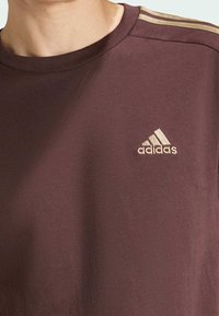 Bordová bavlněná tričko s žebrovaným kulatým výstřihem, krátkými rukávy a zlatými třemi pruhy na ramenou; logo Adidas na levé straně hrudi.