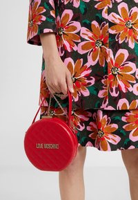 Červená prošívaná kulatá kabelka s řetízkovým uchem, na které je zlaté logo "LOVE MOSCHINO", držena v ruce proti květinovému vzoru oblečení.