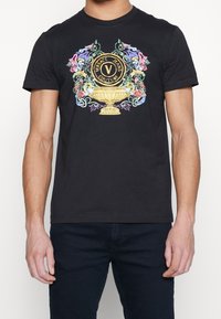 Musta puuvillapaita, jossa on värikäs Versace-logo kukka-akkentteineen ja koristeellinen kultainen uurnakuvio edessä. Lyhyet hihat.