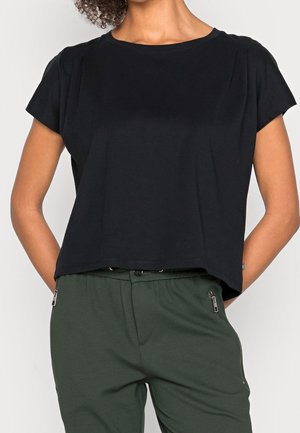 Kobieta ubrana w czarną bluzkę z krótkim rękawem i ciemnozielone spodnie z przednimi kieszeniami na zamek, z rękami założonymi za plecami, na białym tle.