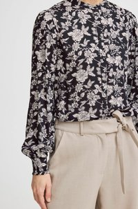 Blusa nera con motivo floreale bianco, caratterizzata da un collo alto e chiusura a bottoni, abbinata a pantaloni chiari beige a vita alta con cintura da annodare.
