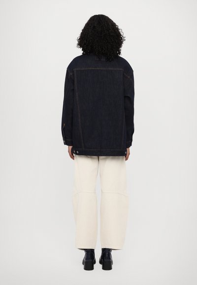 Donkerdenim oversized jas met oranje stiksels, met knopenmanchetten, en een recht gesneden zoom, gedragen over crèmekleurige wijde broekspijpen.