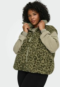 Krøllete person iført grønn fleecevest med leopardmønster over lys grønn genser, holder i kragen nær haken, står foran ensfarget bakgrunn.