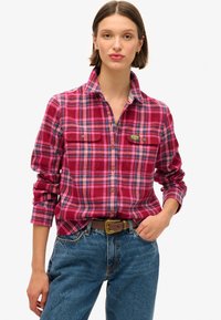 Chemise en flanelle à carreaux rouges et roses, avec un col boutonné, deux poches sur la poitrine et une étiquette de marque, associée à un jean en denim bleu.