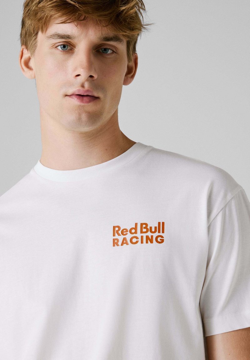 Biała bawełniana koszulka z krótkim rękawem, z małym pomarańczowym logo "Red Bull RACING" na lewej piersi, z okrągłym dekoltem.
