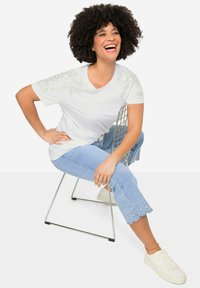 Weiße kurzärmlige T-Shirt mit dekorativen Blumenverzierungen an den Schultern, kombiniert mit hellblauen, cropped Jeans mit Spitzenbesatz und weißen Sneakers.