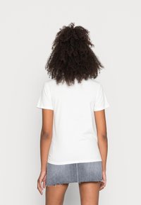 Žena s kudrnatými vlasy, oblečená v jednoduchém bílém tričku s krátkým rukávem a roztrhané džínové mini sukni, pohledem zezadu.