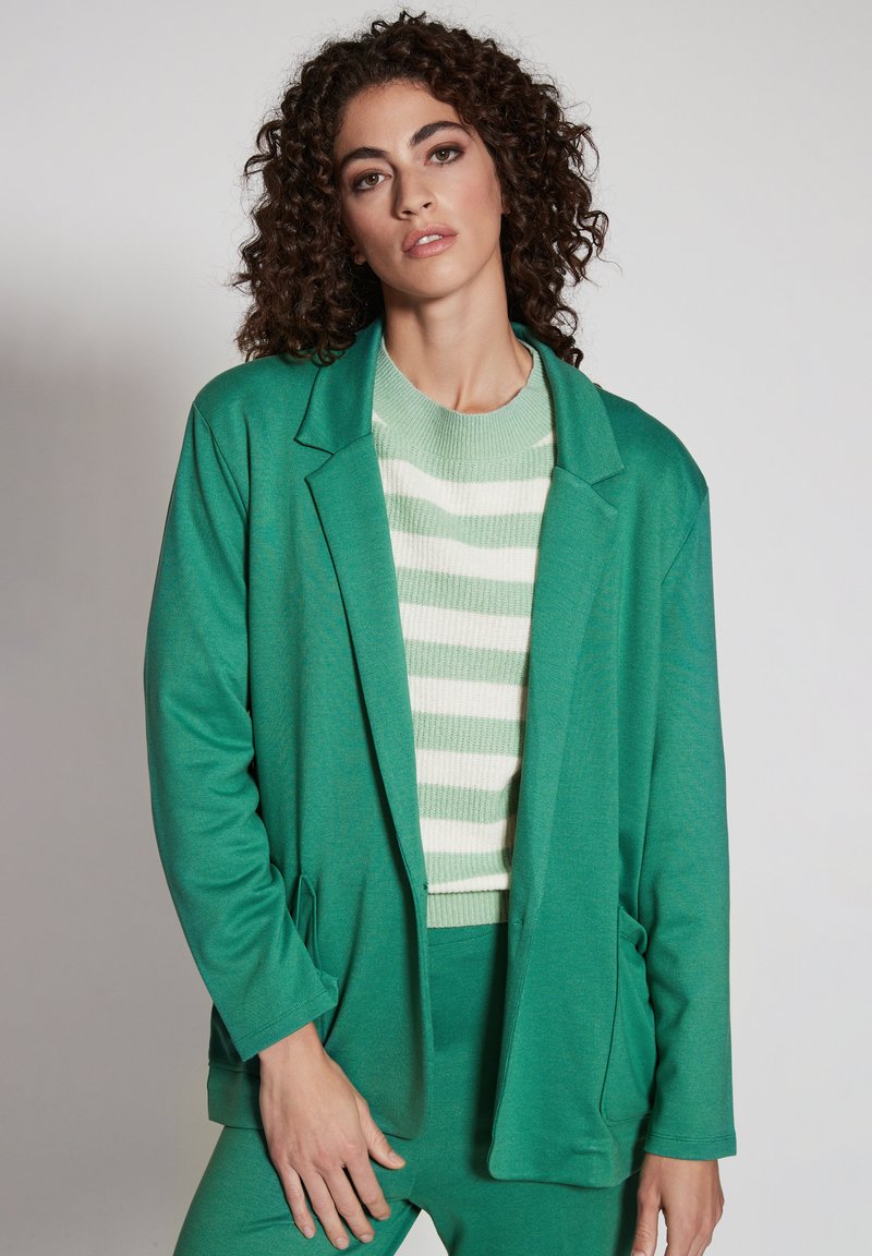 Turkos kavaj med avslappnad passform, långa ärmar och två framfickor, som bärs över en grön och vit randig stickad tröja.