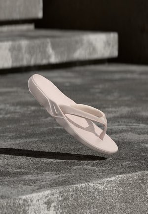 Sandalia tipo chancla Nike de color rosa claro flotando sobre una superficie de hormigón con escalones de piedra bajo una luz solar suave.