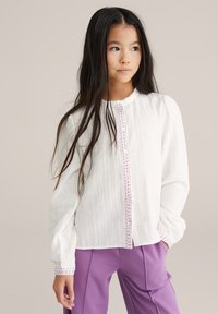 Bílá košile s dlouhým rukávem z texturované látky, s volánkovým výstřihem a fialovými knoflíkovými detaily. V páru s fialovými kalhotami.