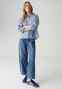 Chemise rayée bleu et blanc à boutons, jean en denim bleu coupe ample, et baskets bleu marine avec des rayures blanches. Tenue décontractée ; silhouette décontractée.