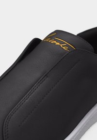 Μαύρο παπούτσι slip-on με λείο δερμάτινο επάνω μέρος, εμφανείς ραφές, λογότυπο σε χρυσό γραφικό στο γλώσσα και λευκή λαστιχένια σόλα.