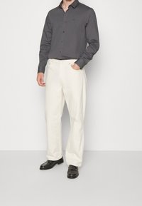Man in een donkergrijs overhemd met knopen, off-white rechte broek en zwarte leren instappers, staande met één hand in zijn zak.