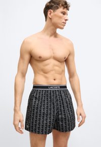 Homme torse nu portant un boxer Lacoste noir à motifs, regardant vers la droite sur un fond clair uni.