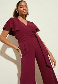 Burgundių jumpsuit'as su trumpomis plačiomis rankovėmis, apvyniojimo dizainu, rišamu liemeniu ir šoninėmis kišenėmis. Lygus audinys su subtiliu blizgesiu. Auksinė apyrankė ant riešo.