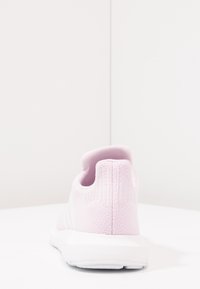Světle růžová pletená sportovní obuv viděná z zadní strany, ukazující texturovanou patu a bílou podrážku na bílé ploše s bílým pozadím.