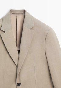 Beige colbert met enkele rij knopen, met revers met inkeping en één zwarte knoop, plat weergegeven op een witte achtergrond.