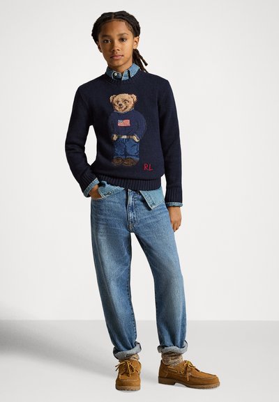 Ung person med flettehår iført en marineblå genser med bamsemotiv, denimskjorte, løse jeans, mønstrede sokker og brune sko.