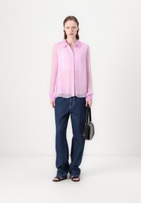 Helles rosa Chiffon-Blusenoberteil mit rüschenbesetztem Kragen, langen transparenten Ärmeln und gerafften Manschetten, kombiniert mit lockeren dunkelblauen Jeans und einer schwarzen Handtasche.