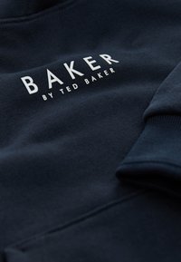 Marineblå sweatshirt med blød tekstur, med "BAKER by TED BAKER" i hvidt tryk; nærbillede der viser detaljer ved ærmekanten og syninger.