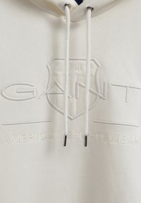 Sudadera color crema con logo y texto "GANT" en relieve. Presenta cordones con puntas de metal, textura de tela suave y un diseño deportivo.