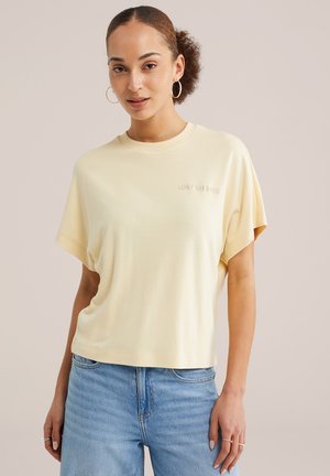 Mlada ženska s kodrastimi lasmi, ki nosi velike obroče, svetlo rumeno ohlapno majico in svetlo modre kavbojke, pozira na nevtralnem ozadju.