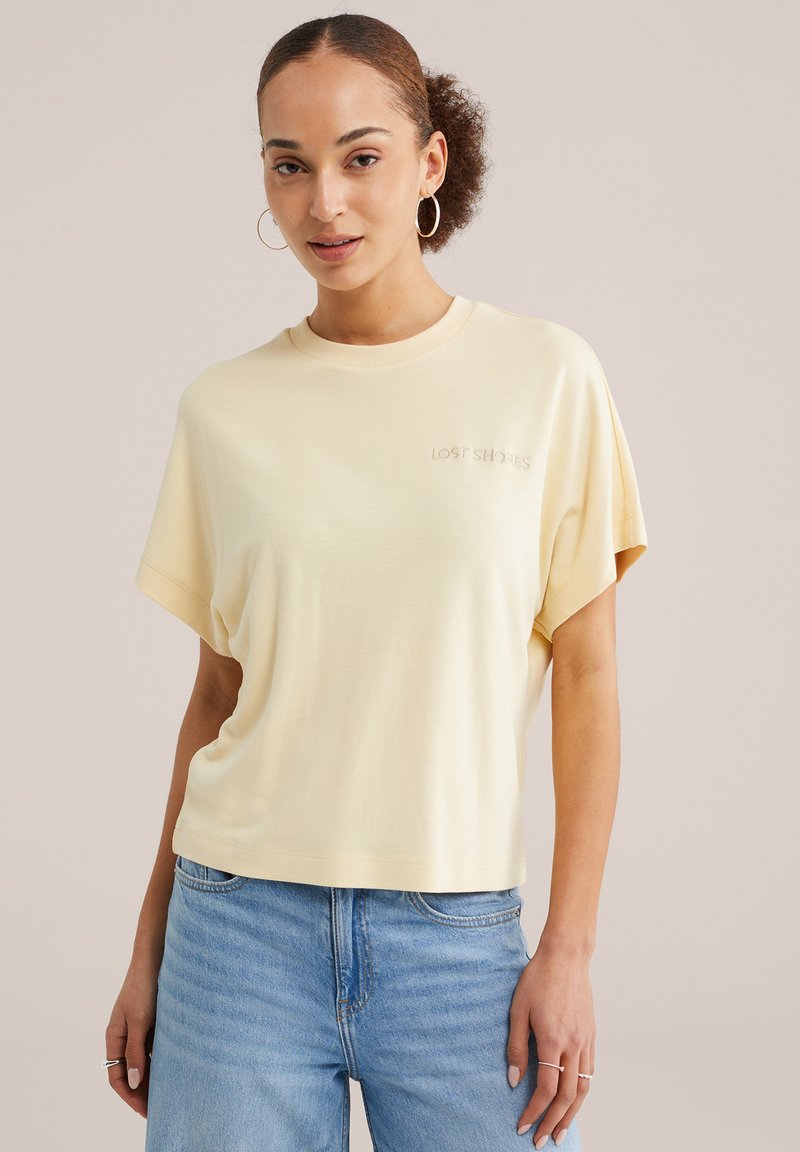 Jeune femme aux cheveux bouclés portant des boucles d'oreilles créoles, un t-shirt ample jaune pâle et un jean bleu clair, posant devant un fond neutre.