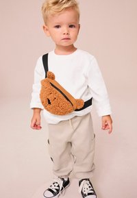 Brun nallebjörnsformad crossbody-väska med svarta detaljer, texturerat tyg, bärs över en vit långärmad tröja och beige byxor, kombinerad med svarta sneakers.