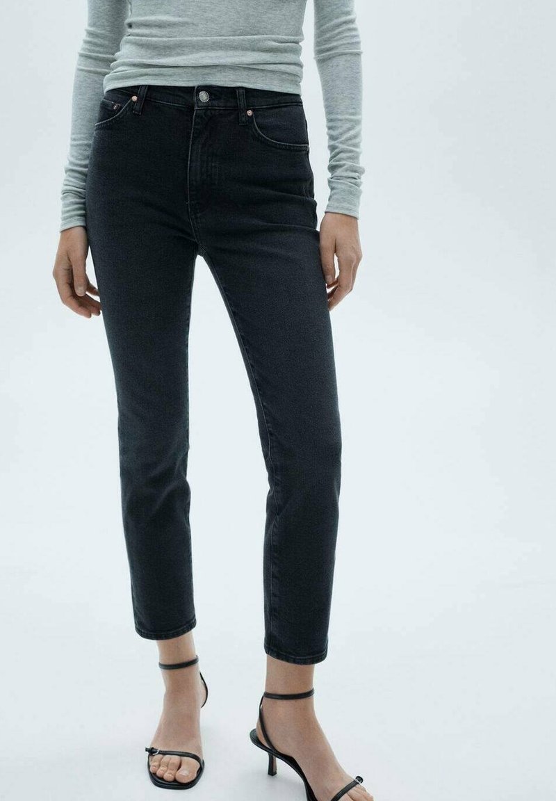 Femme portant un jean noir slim, un haut à manches longues gris clair et des sandales noires à talons hauts et à brides, debout devant un fond uni.