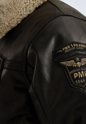 Veste en cuir noir avec un col en fausse fourrure, présentant une surface texturée et un patch avec des ailes et un logo.
