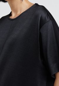 Svart kortärmad topp med rund halsringning, slät textur och subtil lyster, med axeldetaljer för ökad designintresse.