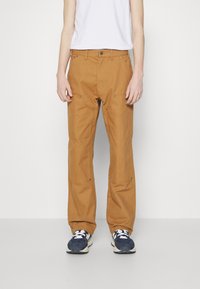 Brune cargo-stil bukser med lige pasform, der har lommer og metal detaljer; båret med en hvid t-shirt og blå sneakers.