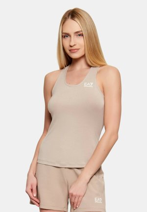 Donna che indossa un top beige senza maniche e short abbinati con logo EA7 Emporio Armani, in piedi davanti a uno sfondo chiaro e uniforme.