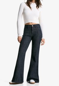 Tmavě modré rozšířené džíny s vysokým pasem, vybavené dvěma předními kapsami a jedním knoflíkem na zapínání, doplněné bílým žebrovaným crop topem.