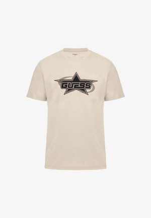 Beige kokvilnas t-krekls ar melnu zvaigžņu grafiku un vārdu "GUESS" treknajiem burtiem. Īsas piedurknes, apaļa kakla izgriezums un gluda tekstūra.