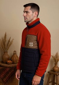 Fleecejack met een rode bovenkant, marineblauwe onderkant en een bruine borstzak met logo. Voorzien van een ritssluiting en een gestructureerde stof.