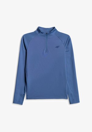 Enfant souriant debout, portant un haut bleu à manches longues zippé, un pantalon assorti et des baskets blanches sur un fond gris uni.