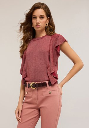Una donna con capelli ondulati indossa un top rosso luccicante con maniche a volant, pantaloni rosa con tasche con bottoni e una cintura scura con fibbia dorata.